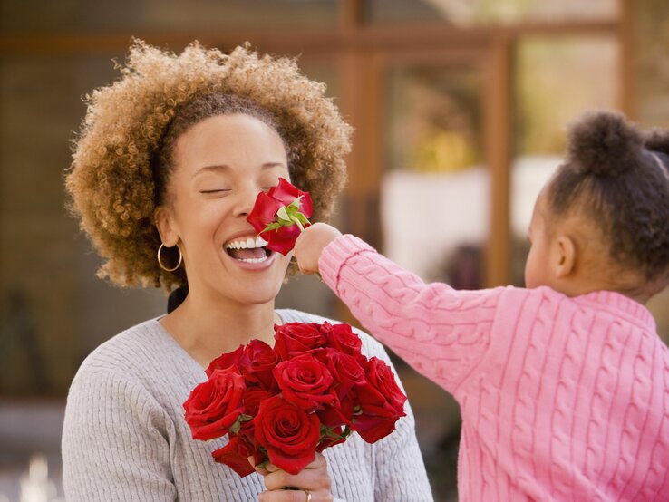 Fröhliche Szene einer Mutter, die eine Rose schnuppert, gereicht von ihrer kleinen Tochter im rosa Pullover.