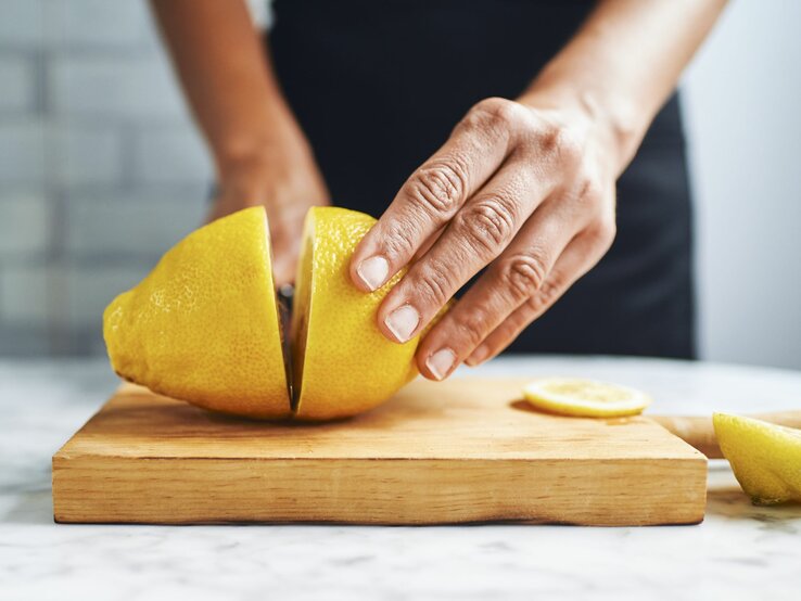 Eine Person mit dunkler Haut schneidet eine Zitrone auf einem Holzbrett in einer hellen Küche.