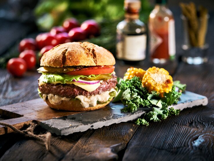 Ein veganer Elote-Burger mit Gemüse, weißer Maissoße, daneben liegen geschnittene Maiskolben und Salatblätter, im Hintergrund sieht man unscharf weitere Zutaten.