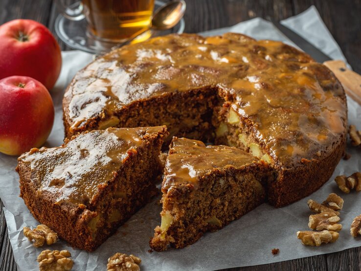 Glasierter Apfel-Walnusskuchen auf hölzernem Untergrund umgeben von den Hauptzutaten, einer Tasse und einem Messer. 