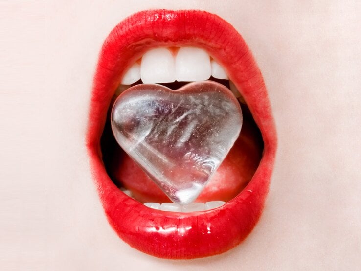 Close-up von roten Lippen. Die Zähne halten einen Eiswürfen in Herzform. 