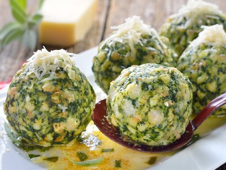 Fünf Spinatknödel auf einem länglichen weißen Teller mit der Salbeibutter. Im Hintergrund ein rot-weiß-kariertes Tuch und ein Stück ganzer Parmesan.