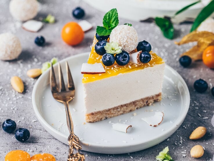 Ein Stück Kokos-Käsekuchen auf einem Teller mit einer Kuchengabel. Drumherum verstreut liegen Physalis, Kokosbällchen, Blaubeeren und Kokosraspeln.