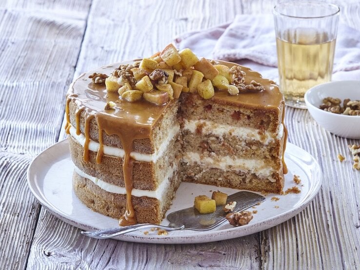 Auf einem Teller steht eine Apfel-Karamell-Schichttorte. Sie ist mit Äpfeln und Nüssen bedeckt. 