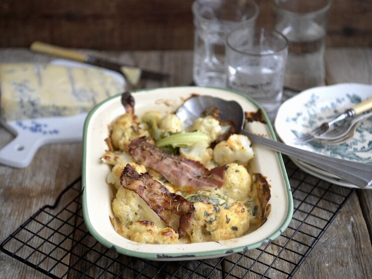 Blumenkohlgratin mit Blauschimmelkaese und gerösteten Speckstreifen, serviert in einer Auflaufform auf einem Rost auf einem gedeckten Holztisch. 