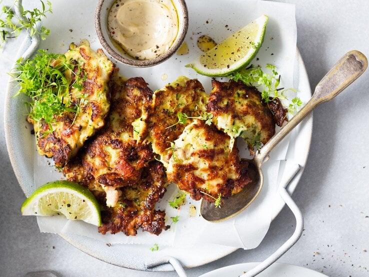 Mehrere Hüttenkäse-Taler mit Zucchini auf einem weißen Teller mit Limette, Kresse und einem hellen Dip auf. Alles auf einem hellen Tisch, direkt von oben fotografiert.