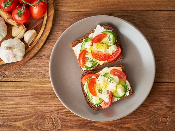 Draufsicht: Zwei Scheiben Brot mit Frischkäse, Gurken- und Tomatenscheiben und Ei liegen auf einem runden Teller. Der Teller steht auf einem Untergund aus Holz. Links daneben ist ein rundes Holzbrett teilweise zu sehen. Auf dem liegen Tomaten und Knoblauch.