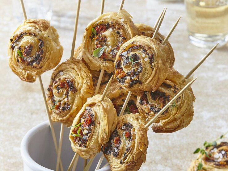 In einem Glas stehen mehrere Blätterteig-Parmesan-Lollies. Sie sind knusprig und goldbraun gebacken. 