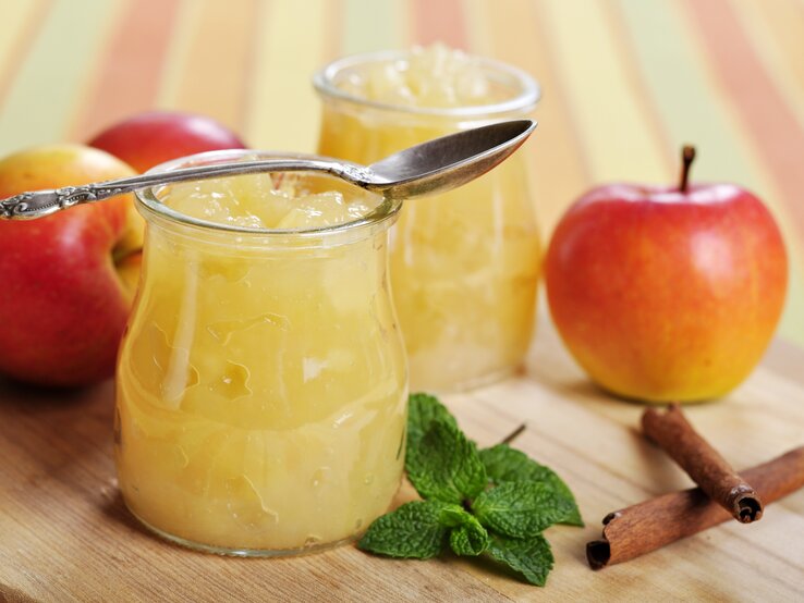 Zwei Gläser Apfel-Minz-Marmelade stehen auf einem Holzbrett. Drumherum liegen Äpfel.