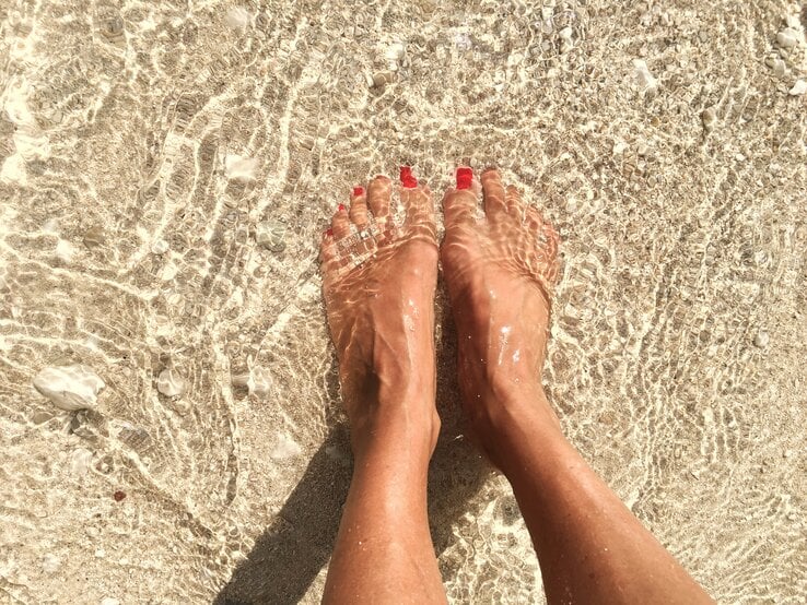 Füße einer Person, die im klaren, flachen Meerwasser am Strand stehen. Die Zehen sind mit rotem Nagellack lackiert. Man sieht, wie das Sonnenlicht das Wasser auf dem sandigen Untergrund funkeln lässt.