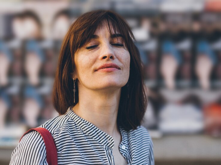 Frau mit geschlossenen Augen und einem entspannten Ausdruck, die draußen vor einem unscharfen Hintergrund steht. | © iStock/filadendron