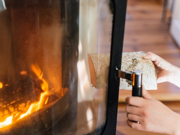 Person öffnet Kaminscheibe und wirft Holz in das Feuer des Kamins.