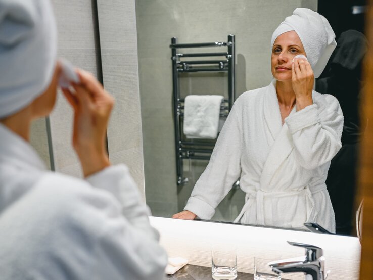 Frau mittleren Alters in einem Badezimmer, die sich selbst im Spiegel betrachtet, während sie ihr Gesicht mit einem Wattepad reinigt. Sie trägt einen weißen Bademantel und ein Handtuch um den Kopf gewickelt, was auf eine Pflegeroutine hindeutet.