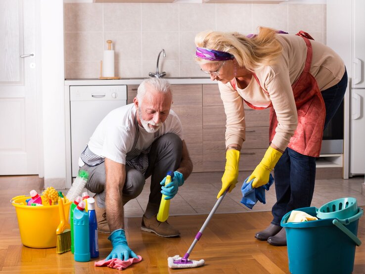 Älteres Paar, das gemeinsam den Fußboden in der Küche putzt. Beide tragen Gummihandschuhe und arbeiten mit verschiedenen Reinigungsmitteln. Der Mann kniet auf dem Boden und benutzt einen Lappen, während die Frau einen Wischmopp verwendet.