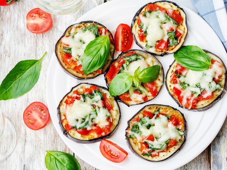 Mehrere Auberginen-Pizzen auf einem weißen Teller und hellem Holztisch von oben fotografiert. | © Shutterstock/ Nataliya Arzamasova