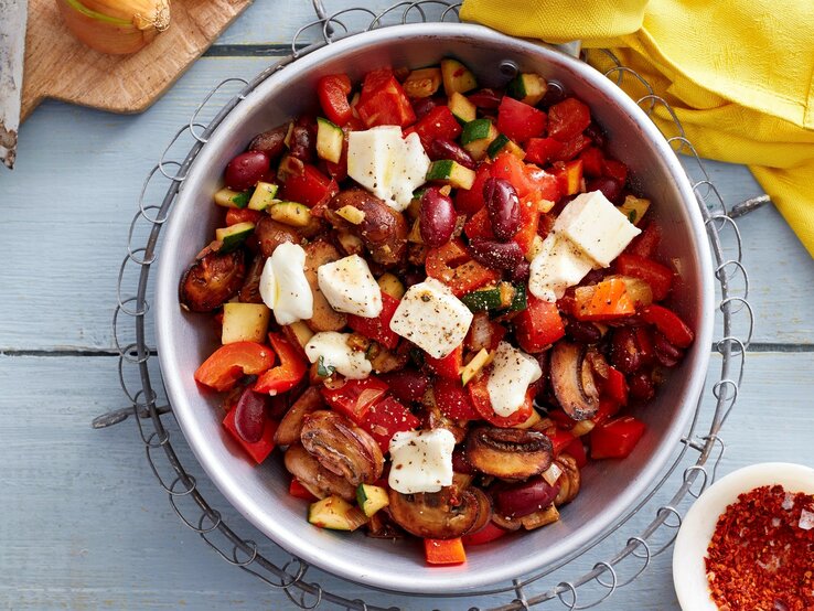 Eine Schüssel Gemüse-Mozzarella-Pfanne in der Draufsicht vor hellem Hintergrund, Chiliflocken, Zwiebel und ein gelbes Küchentuch drum herum. 