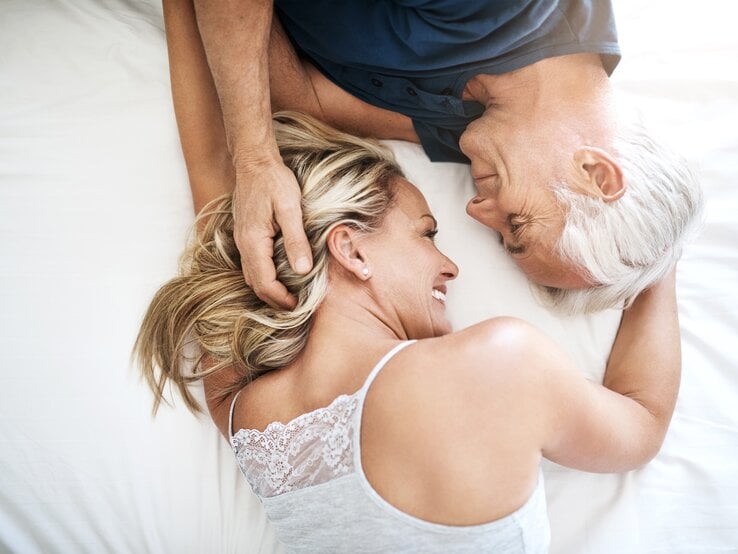 Älteres Paar, das auf einem Bett liegt und sich liebevoll anschaut. Der Mann, mit grauem Haar, streichelt sanft das Haar der Frau, die ein breites Lächeln zeigt. 