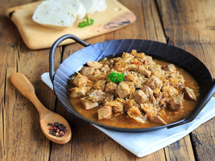 Frontalsicht: Eine schwarze, gusseiserne Pfanne mit Szegediner Gulasch liegt auf einem weißen Tuch, dass auf einem Holztisch liegt. Daneben liegen ein Holzlöffel mit Pfefferkörnernn. Und im Hintergrund sind aufgeschnittene Knödel.