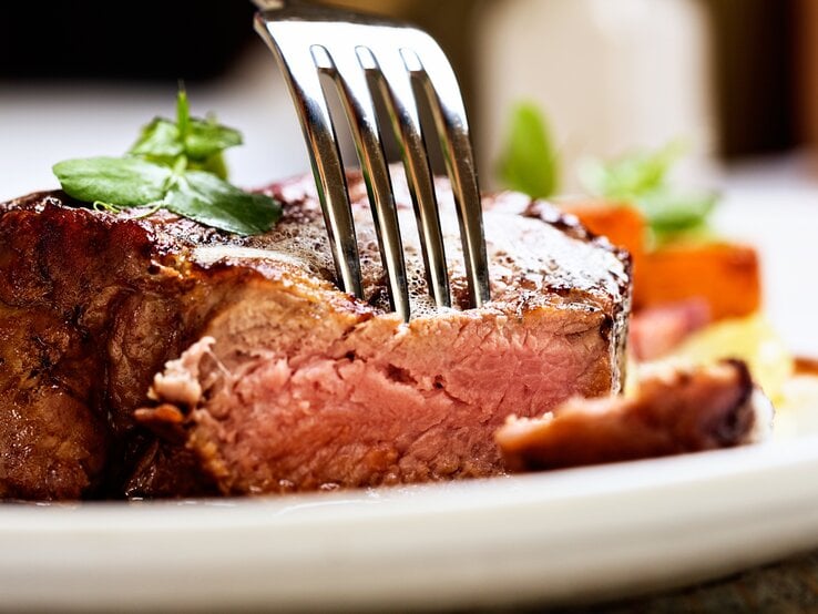 Gabel steckt in einem saftigen Stück Fleisch auf weißem Teller.