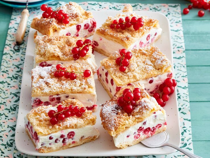 Sechs viereckige Johannisbeer-Knusperschnitten mit frischen Johannisbeeren auf einem rosa Teller auf einem gemusterten Tuch mit blauem Holzuntergrund. 