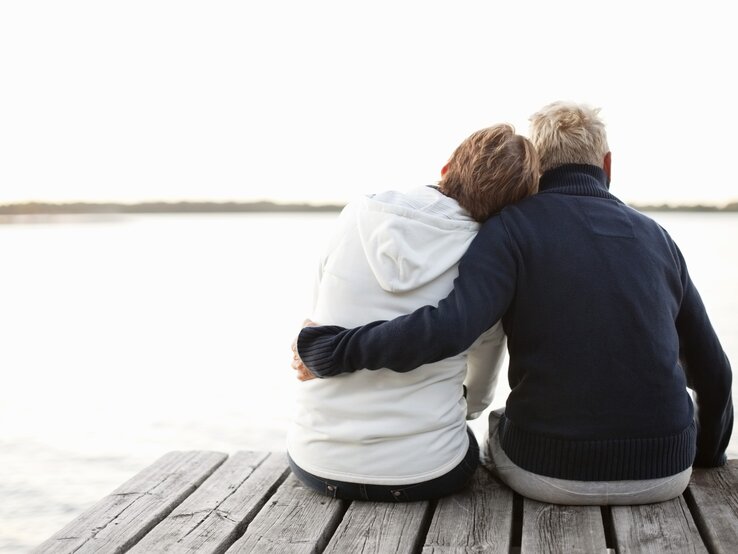 Zwei Personen, die auf einem Steg sitzen und aufs Wasser blicken. Sie sind von hinten zu sehen und tragen warme Kleidung, vermutlich Pullover oder Jacken. Eine Person hat den Arm um die andere gelegt.