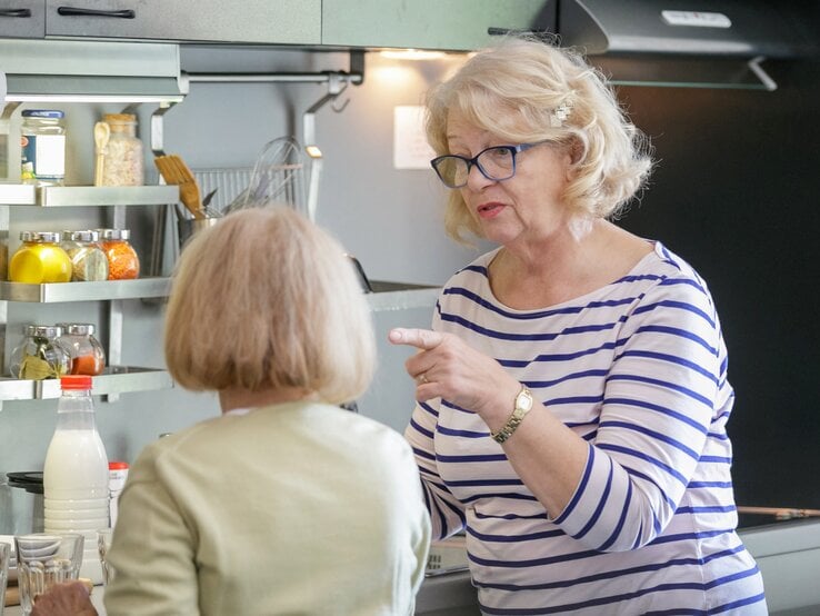 Zwei ältere Frauen sind in einer Küche in eine Unterhaltung vertieft. Die Frau im Vordergrund, mit blonden Haaren und Brille, gestikuliert mit ihrem Finger, was auf eine ernsthafte Diskussion oder Erklärung hinweist.