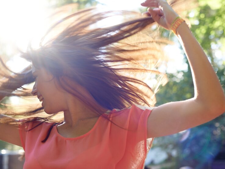 Eine junge Frau mit langen braunen Haaren tanzt lachend in der Sonne, ihr rosafarbenes Oberteil leuchtet im Gegenlicht.