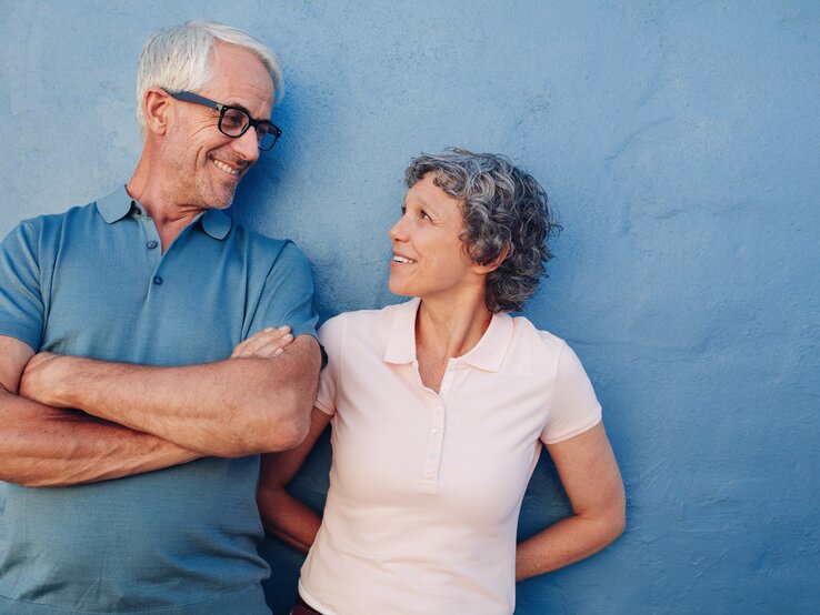 Ein älteres Paar mit grauem Haar steht lachend vor einer blauen Wand, der Mann trägt ein blaues Poloshirt, die Frau ein rosafarbenes.