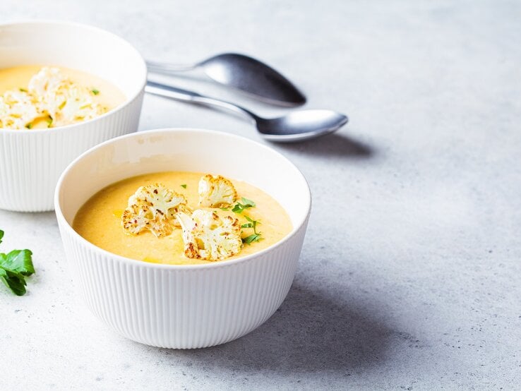 Zwei Schüsseln Blumenkohl-Kartoffel-Suppe mit Kräutern und gerösteten Blumenkohlröschen garniert, dahinter zwei Löffel.