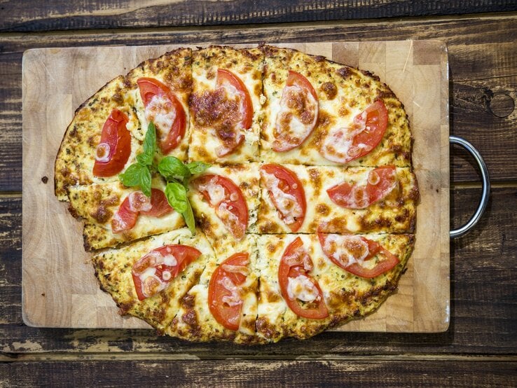 Mit Tomaten belegte Pizza auf Holzbrett, drapiert auf hölzernem Untergrund. 