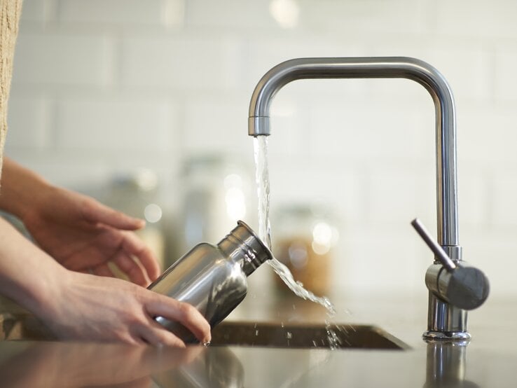 Person füllt eine Wasserflasche aus Edelstahl an einem Wasserhahn auf. Die Szene spielt sich in einer modernen Küche ab, die eine saubere, helle und minimalistische Atmosphäre hat.