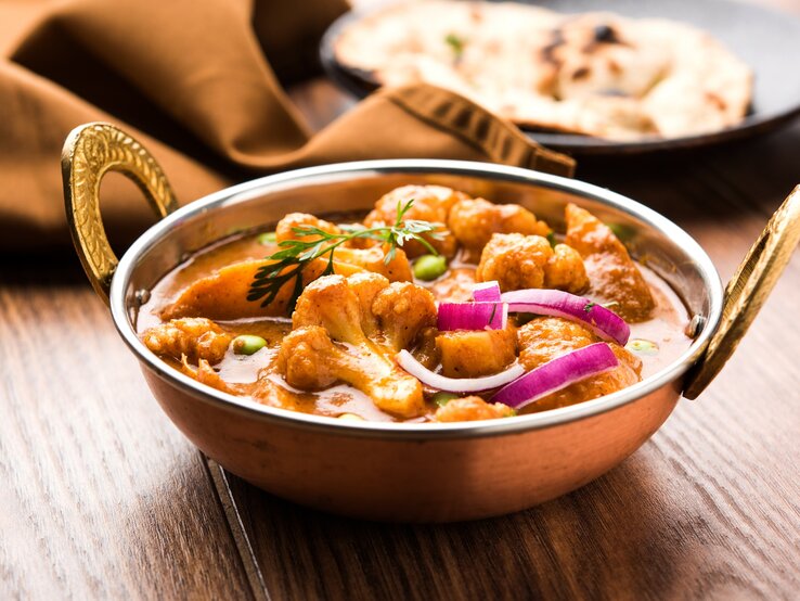 Eine Schüssel Aloo Gobi, ein Kartoffel-Blumenkohl-Curry, mit roter Zwiebel und Kräutern, im Hintergrund ein Naan-Brot.