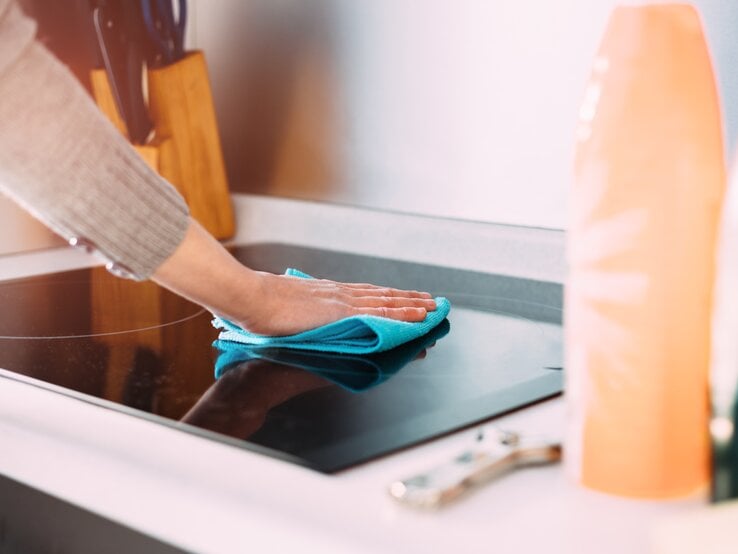 Person, die eine Küchenarbeitsplatte mit einem blauen Mikrofasertuch reinigt. Der Fokus liegt auf der Hand der Person, die das Tuch über die glänzende, schwarze Oberfläche eines Kochfeldes führt.