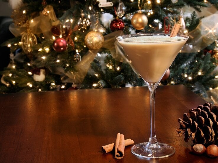 Eierlikör-Martini im Glas mit Zimtstange auf Holztisch mit Zimtstange, Haselnüssen und Tannenzapfen. Im Hintergrund Weihnachtsdekoration. Frontalansicht.