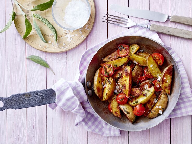 Eine gusseiserne Pfanne mit mediterranen Kartoffeln drinnen, die auf einem rosa karierten Geschirrtuch liegt neben Besteck, Salbei und grobem Meersalz