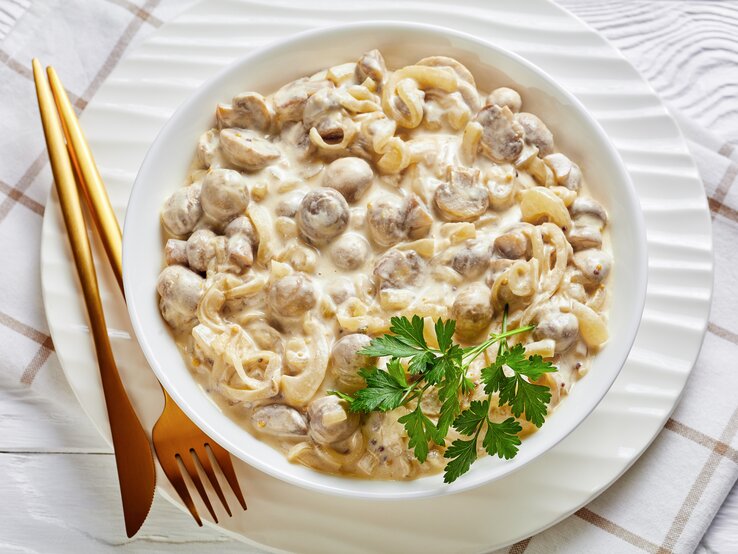 Rahmchampignons mit Nudeln auf weißem Teller, angerichtet auf einem hellen Tischtuch und zu Holzbesteck.