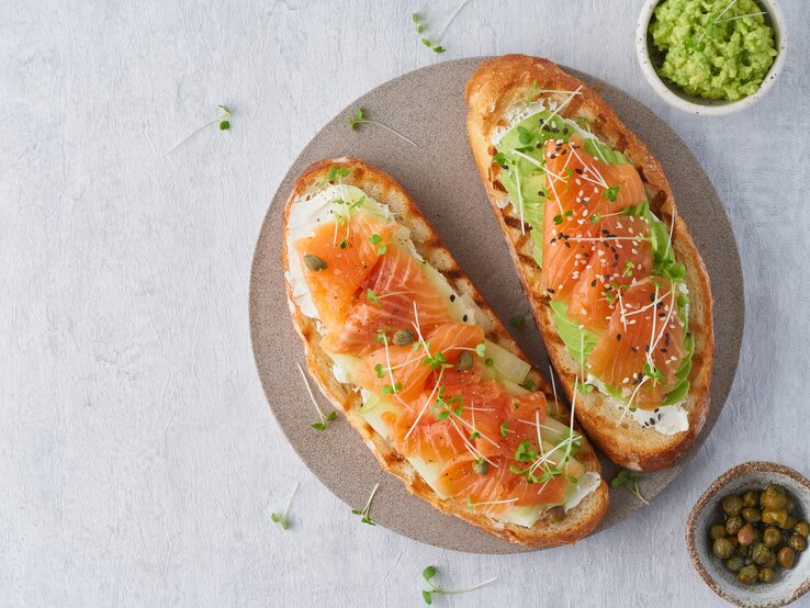 Draufsicht: Ein Teller auf dem zwei Low-Carb-Toast mit Lachs liegen. Daneben ist ein rundes Gefäß mit Avocadocreme und eines mit Kapern.