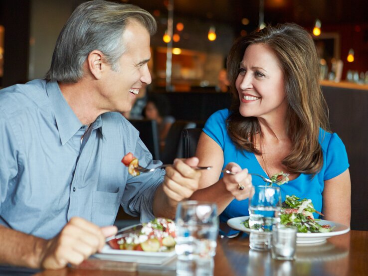 Älteres Paar beim Essen in einem Restaurant. Der Mann, mit grauem Haar und einem hellgrauen Hemd, sitzt seiner Partnerin gegenüber und lächelt sie an, während er mit einer Gabel Essen zu ihr führt.  | © iStock/monkeybusinessimages