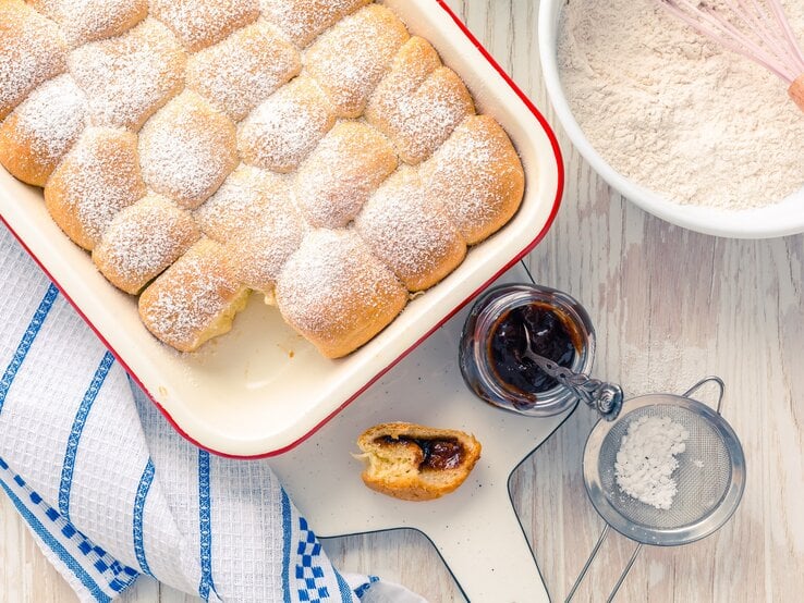 Draufsicht: Weißer Untergrung, auf einem Kuchenhandtuch eine Auflaufform mit selbst gemachten Hefe-Buchteln. Daneben eine Schüssel mit Mehl, ein geöffnetes Glas Fruchtaufstrich, ein Sieb mit Puderzucker und eine angebissene Buchtel.