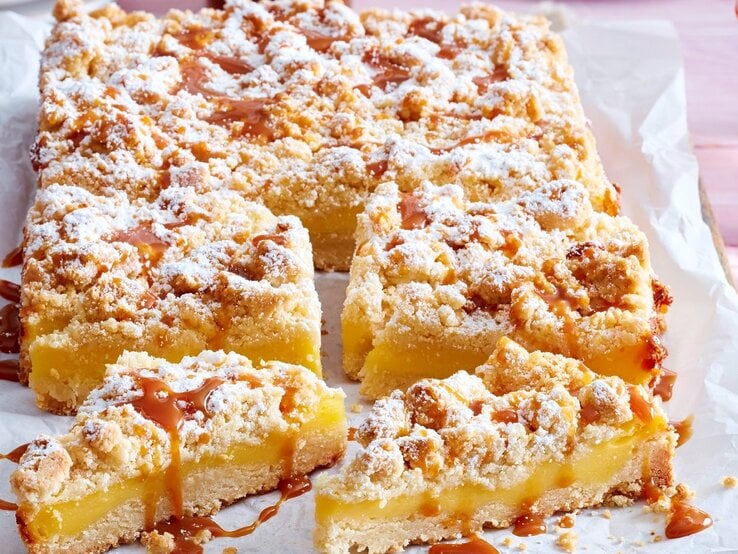 Auf einem Backblech liegt ein Streusel-Puddingkuchen. Er ist bestreut mit Puderzucker und mit Karamellsoße überzogen. Man erkennt die cremige Pudding-Füllung. 