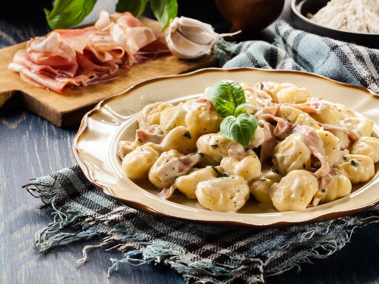 Gnocchi in Schinken-Rahm-Soße auf kitschigem Teller, daneben ein Holzbrett mit Schinken und Knoblauch. 