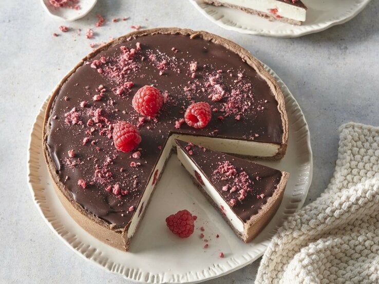 Auf einem Teller steht ein Käsekuchen mit Schokolade und Himbeeren. Er ist mit Himbeeren und Schokolade überzogen. 