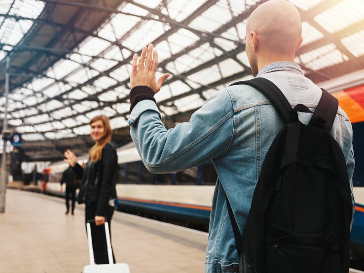 Zwei Personen verabschieden sich an einem Bahnhof voneinander. Im Vordergrund steht eine Person mit dem Rücken zur Kamera, die eine Jeansjacke und einen schwarzen Rucksack trägt und der anderen Person zuwinkt. Die zweite Person steht weiter hinten auf dem Bahnsteig, ebenfalls winkend, und hält einen Koffer. Im Hintergrund ist ein Zug zu sehen.