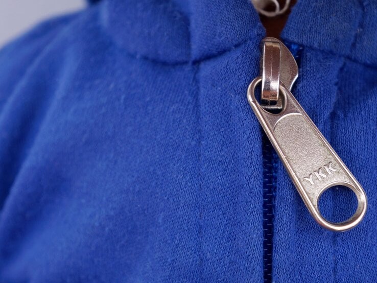 Nahaufnahme eines Reißverschlusses an einem blauen Kleidungsstück. Auf dem silbernen Zipper ist der Schriftzug "YKK" eingraviert.