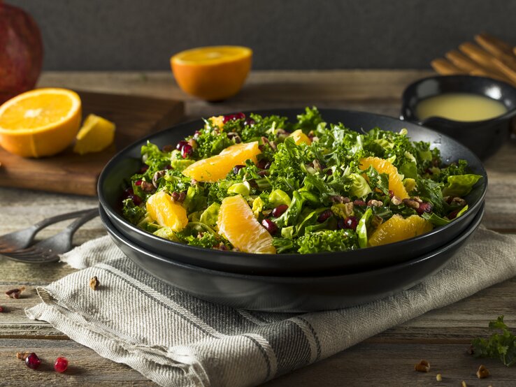 Auf einem Tisch steht ein schwarzer Teller. In dem ist ein Orangen-Grünkohl-Salat. Auf dem Tisch liegen Orangen-Hälften und ein Granatapfel. | © Shutterstock / Brent Hofacker