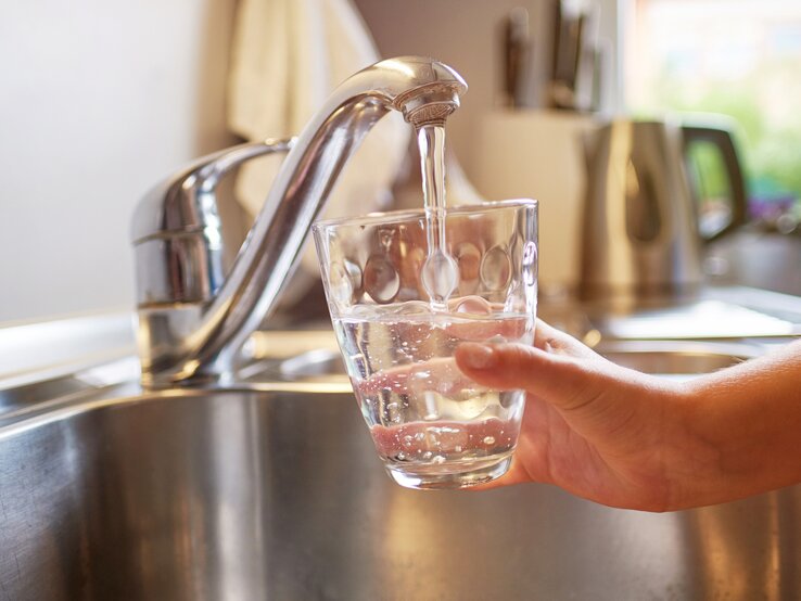 Das Bild zeigt eine Person, die ein Glas mit klarem Wasser aus einem modernen, metallischen Wasserhahn in einer Küchenspüle füllt. Das Tageslicht fällt durch ein Fenster ein und beleuchtet die Szene, was auf eine häusliche und gesunde Umgebung hinweist. Das Wasser sieht sauber aus und die Szene kommuniziert die Zugänglichkeit von sauberem Trinkwasser, was für eine gesunde Lebensweise unerlässlich ist.