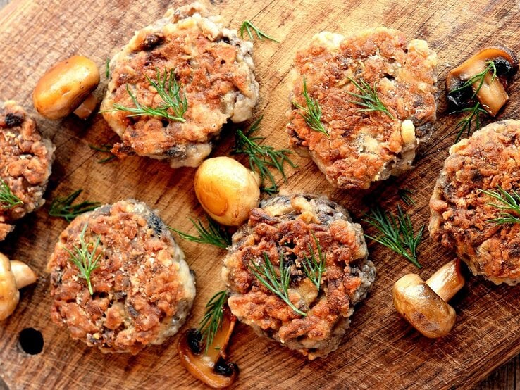 Mehrere Champignon-Bouletten auf einem Holzbrett mit gebratenen Pilzen und Kräutern.