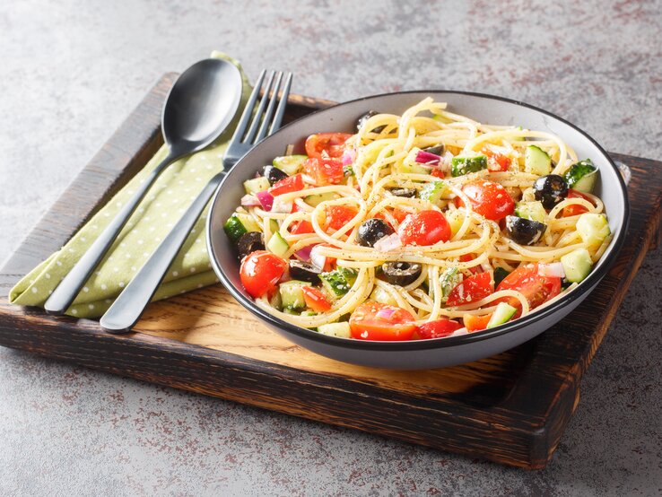 Kalifornischer Nudelsalat mit Zucchini, Paprika, Tomate, Zwiebel, Gurke und Oliven in einer Schale, angerichtet auf einem Holzbrett. Daneben liegt Besteck. 