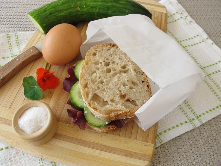 Ein belegtes Brot mit Butterbrottüte, ein Ei, eine Gurke, ein Messer und ein Schälchen Salz liegen auf einem Küchenbrett aus Holz.