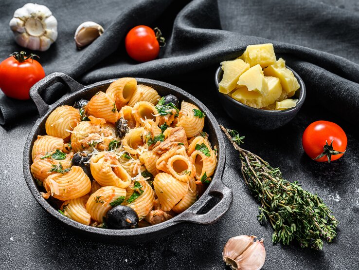 Draufsicht: eine runde schwarze Schüssel Pasta mit Oliven und Tomaten auf schwarzem Untergrund. Drum herum liegen frische Tomaten, frischer Thymian, frischer Knoblaucg und eine Schale mit Parmesanstücken.
